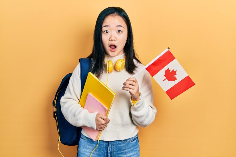 Young chinese girl exchange student holding canada flag afraid and shocked with surprise and amazed expression, fear and excited face